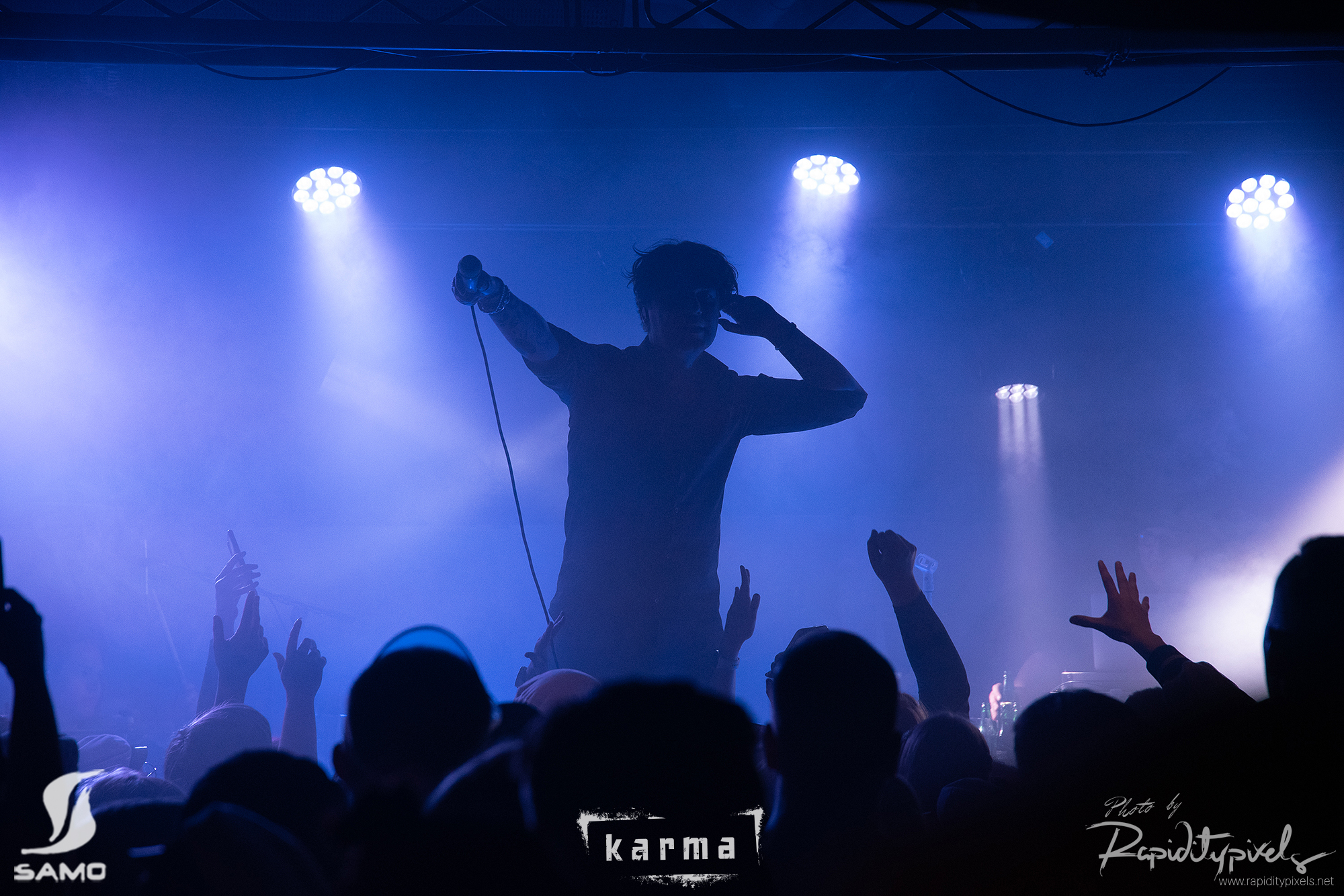 SAMOn Wapu 2023. Siluetti esiintyjästä keskittyneenä mikrofoniin, yleisön kädet ilmaan noustessa ja valokeilan osuessa lavalle.